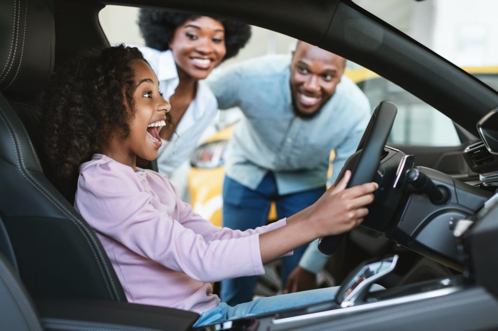 Black girl sitting in driver seat of new car, shouting in excitement, helping her parents buy