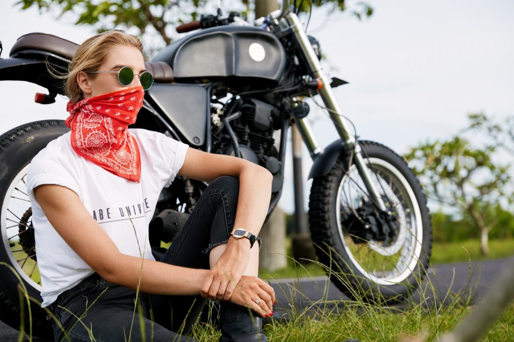 Stylish biker model poses near motorbike, sit on ground outdoor, wears sunglasses and bandana on fac